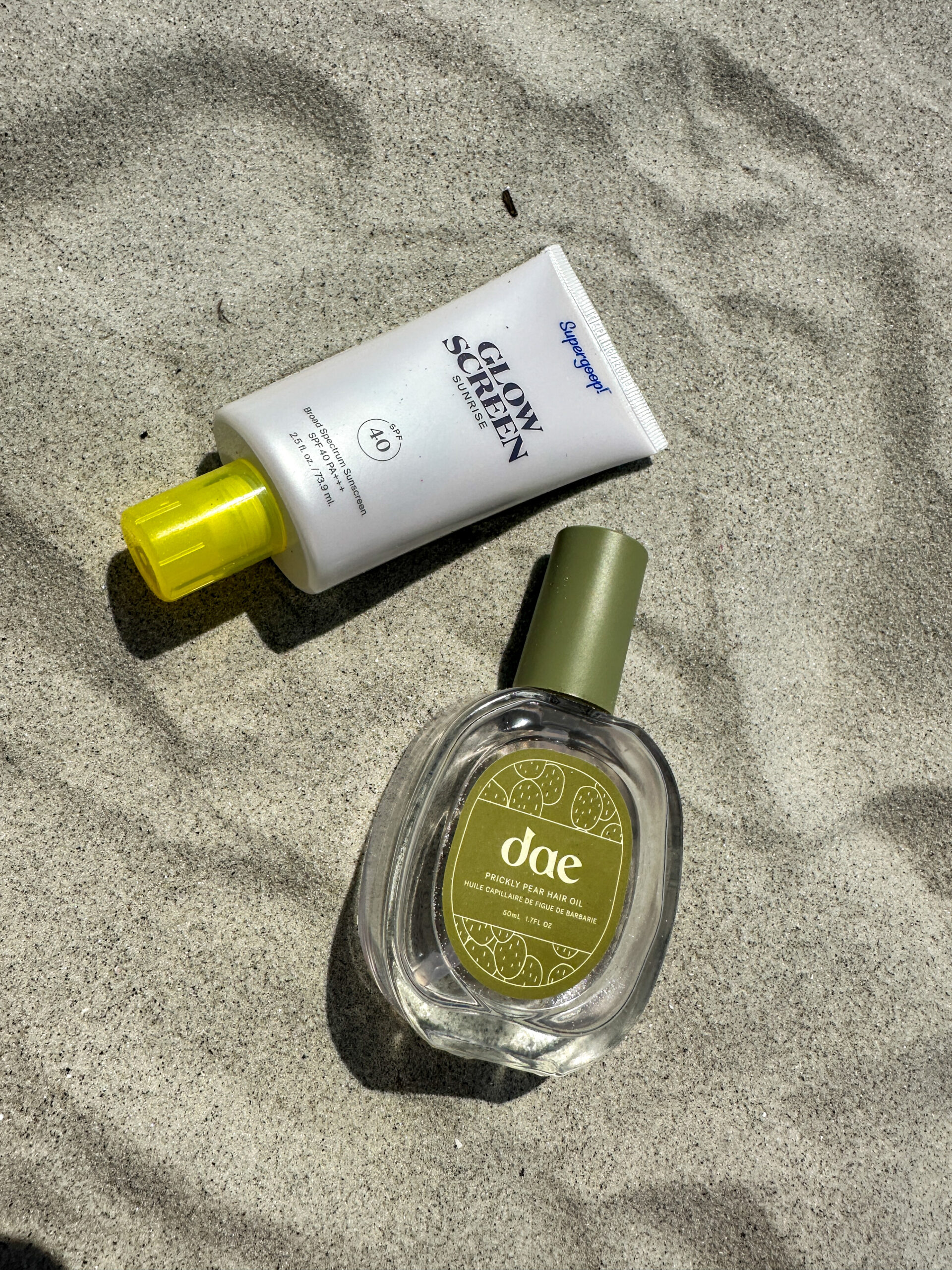 a bottle of superg oop sunscreen and bottle dae hair oil flat laying on the sand